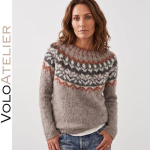 Può includere: Un maglione a maniche lunghe in maglia soffice con scollo rotondo e un motivo fair isle sul petto e sulle spalle nei toni del marrone, grigio, bianco e arancione ruggine. Il maglione è indossato sopra una camicia bianca e jeans blu.