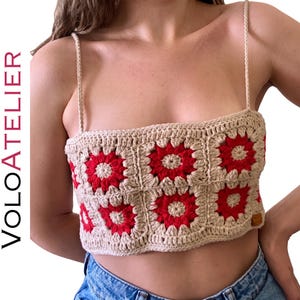 Puede incluir: Un top corto de ganchillo en color beige y rojo con un diseño de cuadrados y motivos florales. La prenda tiene tirantes finos trenzados y un escote recto. El texto VOLOATELIER es visible en el lado izquierdo de la imagen en vertical.