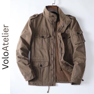 Könnte beinhalten: Eine braune Feldjacke mit Stehkragen, Frontreißverschluss mit Druckknöpfen, vier Vordertaschen mit geknöpften Klappen und einer kleinen Tasche am linken Ärmel. Der Text VoloAtelier ist vertikal auf der linken Seite aufgedruckt.