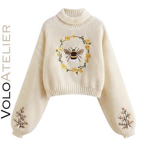 May include: A cream-colored ribbed knit sweater with a mock neck and long sleeves. The front features an embroidered bee centered within a circular wreath of yellow flowers. Additional floral embroidery appears on the lower part of each sleeve. The text VOLOATELIER is printed vertically along the left side.