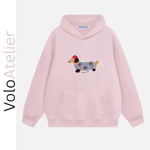 Può includere: Una felpa con cappuccio rosa chiaro caratterizzata da una grafica di un bassotto che indossa un maglione a righe blu e un basco rosso. Il testo VoloAtelier è stampato verticalmente in nero sul lato sinistro. Include una tasca a marsupio.