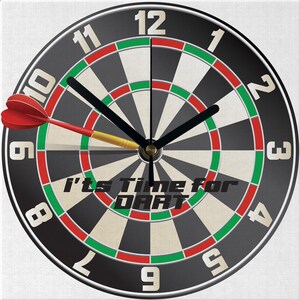 May include: A round wall clock featuring a dartboard design. The clock face is black and white with red and green accents. A red dart is positioned on the board. The text "It's Time for DART" is printed on the clock face.