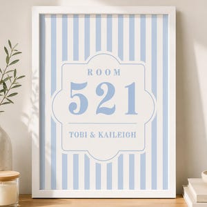May include: A rectangular white frame containing a print with vertical light blue and white stripes. The center features a decorative white label shape with the text ROOM 521 and the names TOBI & KAILEIGH printed in a light blue serif font.