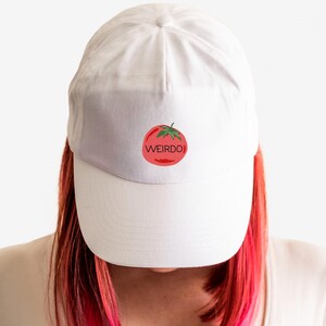 May include: White baseball cap with a red tomato graphic and the word "WEIRDO" in a playful font. The cap is worn by a person with red and pink hair, against a white background. The cap is a casual accessory.