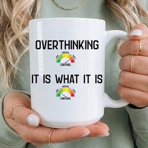 May include: White ceramic mug with the text "OVERTHINKING IT IS WHAT IT IS" in black. The mug features two speedometer graphics. The mug is held by a person wearing a green sweatshirt.