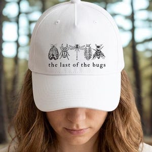 May include: White baseball cap featuring a black insect illustration and the text "the last of the bugs." The cap is made of a soft material and has a curved brim. The background is a blurred forest scene.