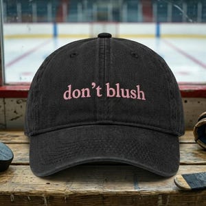 May include: A black baseball cap with a curved brim and an adjustable strap at the back. The front of the cap features the text "don't blush" embroidered in light pink lowercase letters. The cap has a structured crown with ventilation eyelets.