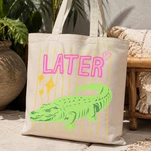 Peut inclure: Un sac fourre-tout en toile beige avec une impression graphique d'un alligator vert vif. Au-dessus de l'alligator, le mot LATER est imprimé en lettres roses style néon, accompagné d'un petit cœur rose et d'étoiles jaunes sur des lignes.
