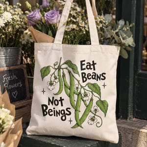 Peut inclure: Un sac fourre-tout en toile couleur crème orné d'une illustration botanique verte représentant des gousses de haricots et des fleurs blanches. Le texte noir imprimé sur le sac indique Eat Beans Not Beings, avec de petites étoiles.