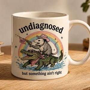 May include: A white ceramic mug featuring a graphic of an opossum in a pink cowboy hat riding a green alligator. The opossum holds a gun with a flag that says HELP. A rainbow arches behind them. Text reads undiagnosed but something ain't right.