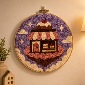 May include: A round, framed textile art piece featuring a whimsical bakery scene. The design includes a shop with a striped awning, cakes, and a cherry on top, set against a purple sky with clouds and stars. The embroidery is in shades of pink, brown, and cream.