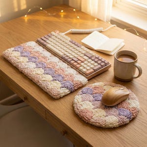 Op de afbeelding: Een houten bureau-opstelling met een toetsenbord, muis en accessoires. Het toetsenbord heeft lichtgekleurde toetsen. Een bijpassende pastelkleurige, getextureerde muismat en toetsenbordpad liggen op het bureau. Een houten muis en een mok koffie maken de scène compleet.