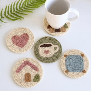 May include: Five round punch needle coasters with cream-colored backgrounds. Designs include a pink heart, a green coaster with a coffee mug, a house with a pink roof, a blue square with brown corners, and one under a white ceramic mug.