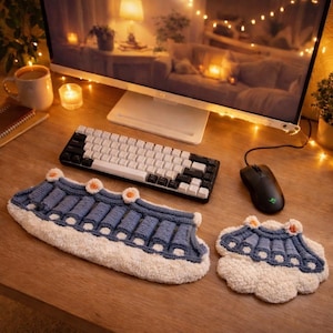 May include: A desk scene with a black and white mechanical keyboard, a black mouse, and a blue and white knitted wrist rest and coaster. The wrist rest and coaster have a distinctive design with orange detailing. A computer monitor and a mug are also present.