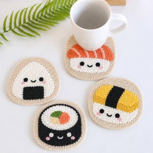 May include: Four round, beige punch needle coasters featuring embroidered sushi designs with smiling faces. One coaster depicts a salmon nigiri, one an onigiri, one a maki roll, and one a tamago nigiri. A white ceramic mug sits on the salmon coaster.