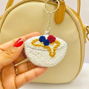 以下が含まれることがあります： 青と赤のベリーのような装飾と金色のシロップのディテールが特徴の、白い刺繍のデザートボウル型のキーホルダー。 キーホルダーはシルバーのキーリングに付けられており、ベージュのハンドバッグを背景にしています。