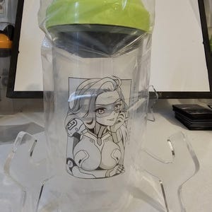 May include: A clear plastic shaker bottle with a lime green lid and an orange flip-top cap. The bottle features a black and white graphic print of an anime-style character with long hair and a visor, enclosed in a rectangular frame on the front.
