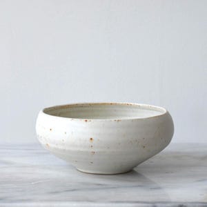 May include: A handmade, off-white ceramic bowl with a rounded form. The bowl has a speckled texture and a slightly flared rim. The interior and exterior have a matte finish. The bowl is on a marble surface.