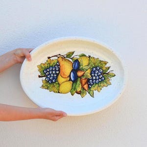 Könnte beinhalten: Ovale Servierplatte aus Keramik mit handgemaltem Fruchtdesign. Das Design zeigt Birnen, Trauben, Zitronen und Pflaumen. Die Platte ist weiß mit einer gesprenkelten Textur und wird von zwei Händen gehalten.