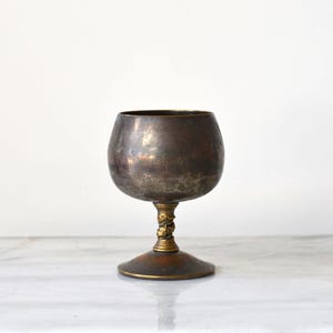 May include: A metallic goblet with a dark, tarnished finish on the bowl and a contrasting gold-toned stem featuring a textured, twisted design. The base is circular and flat, matching the dark, aged patina found on the upper portion of the vessel.