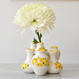 May include: A set of white ceramic bud vases with yellow floral patterns. The vases have yellow rims and hold a large white chrysanthemum. The arrangement is on a marble surface. The vases are in a cluster.