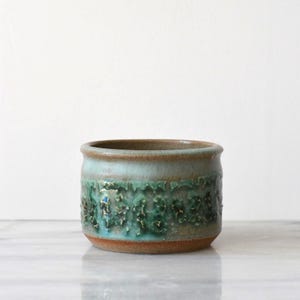 May include: A cylindrical ceramic planter featuring a textured, raised green and turquoise glaze pattern around the middle section. The base and rim are finished in a smooth, earthy brown clay tone, creating a contrast with the vibrant, glossy centre.
