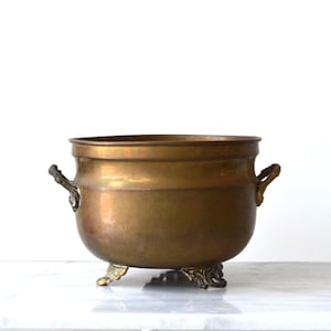 Op de afbeelding: Een ronde koperen plantenbak met een gladde, gepolijste afwerking en een brede, open bovenkant. De pot is voorzien van twee kleine, sierlijke handvatten aan weerszijden en rust op drie gedetailleerde, gebogen voetjes voor een stabiele basis.