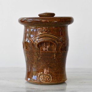 May include: A brown ceramic jar with a lid. The jar has a cylindrical shape with a slightly wider top and a decorative arched design on the front. The jar is glazed with a glossy finish.