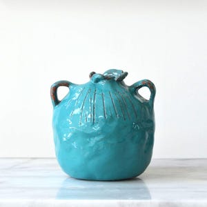 May include: A turquoise ceramic vase with a rounded body, two side handles, and a textured surface featuring etched vertical lines near the top. The glaze has a glossy finish with visible brown accents along the rim and handle edges.