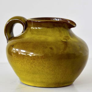 May include: A small, glazed ceramic pitcher in shades of yellow and brown. The pitcher has a rounded body, a curved handle, and a spout for pouring. The glossy finish reflects light, highlighting the color variations.