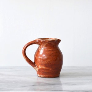 May include: A small, rustic ceramic pitcher with a rounded body and a curved handle. The pitcher is a warm, earthy brown color with lighter streaks and a slightly textured surface. The opening has a scalloped edge.