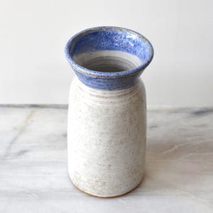 Könnte beinhalten: Eine handgefertigte Keramikvase mit gesprenkeltem weißem Körper und einem ausgestellten Rand. Der Rand ist mit einem blauen Band bemalt. Die Vase hat eine rustikale, organische Ästhetik, geeignet für die Präsentation von Blumen oder als dekoratives Stück.