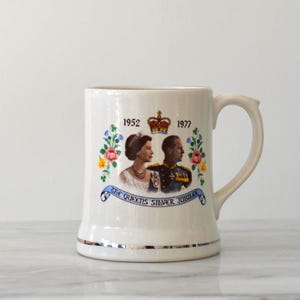 May include: Cream-colored ceramic mug with a handle, featuring a portrait of a royal couple and the text "THE QUEENS SILVER JUBILEE". The mug has a silver band at the base and rim, with floral and crown details.