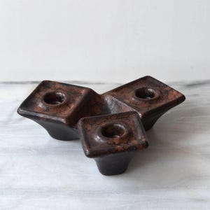 May include: A dark brown ceramic candle holder with a mottled, textured finish. The object features three square-shaped platforms, each with a central circular opening designed to hold a candle, arranged in a triangular, interconnected formation.