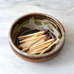 Könnte beinhalten: Eine kleine, runde Keramikschale mit brauner und beiger Glasur, gefüllt mit Holzstreichhölzern. Die Streichhölzer haben braune Spitzen und sind ungeordnet in der Schale angeordnet. Die Schale steht auf einer hellen Oberfläche.
