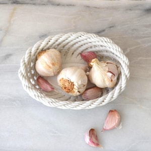 May include: A white, rope-textured basket containing whole garlic bulbs and individual cloves. The garlic is off-white with a hint of brown at the root. The basket is on a marble surface.