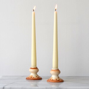 May include: Two tall, slender, cream-colored taper candles are lit, each resting in a ceramic candlestick holder. The holders feature a rounded base with a hand-painted brown floral pattern on a cream background, with a smooth, glossy finish.