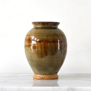 May include: A ceramic vase with a rounded body and a flared rim, featuring a glossy glaze in shades of olive green and brown. The surface displays vertical drip patterns in a darker brown tone, while the base remains unglazed in a light tan colour.