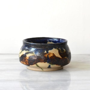 May include: A small, round ceramic bowl with a wide opening and a short, tapered base. The exterior features a mottled, hand-applied glaze with swirling patterns of deep navy blue, dark brown, and cream, with a glossy finish throughout.