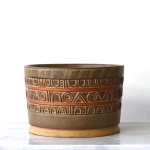 May include: A cylindrical ceramic planter featuring a dark olive-green glaze with a wide, recessed band around the center. This band is decorated with embossed geometric patterns, including arches and circles, highlighted in a burnt orange color.
