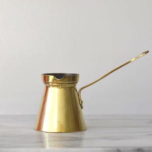 Peut inclure: Une cafetière en laiton doré brillant, avec une longue et fine poignée. Le pot a une base large qui se rétrécit vers le haut. L'intérieur du pot est sombre. Il est posé sur une surface en marbre.