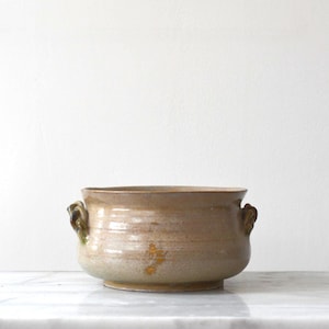 May include: A round ceramic bowl with a wide opening and two small, curved handles on opposite sides. The bowl features a mottled, earthy tan and beige glaze with subtle brown and yellow highlights, showcasing a smooth, glossy finish throughout.
