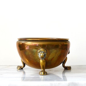 May include: A round, polished copper bowl featuring a smooth, reflective surface and a wide, open top. The bowl is supported by three distinct metal feet, each intricately sculpted in the shape of a lion head with extended paws.