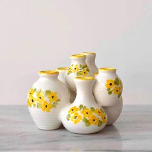 May include: A cluster of six white ceramic vases with yellow rims and hand-painted yellow and green floral designs. The vases are arranged together, creating a unique decorative piece. The background is a light, neutral color.