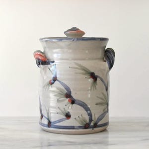 May include: A ceramic jar with a lid, featuring a hand-painted design of bamboo stalks with red and green accents on a cream-colored background. The jar has two handles and a blue rim. The lid is topped with a small, striped knob.