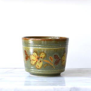 Pode incluir: Um vaso cilíndrico de cerâmica com um esmalte verde-oliva brilhante. O exterior apresenta um padrão floral pintado à mão em tons de amarelo mostarda, laranja queimado e castanho escuro, com uma borda castanha lisa e sulcos horizontais.