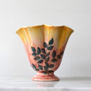 May include: A ceramic vase with a flared, scalloped rim and a pedestal base. The exterior features a gradient glaze transitioning from yellow at the top to a reddish-pink hue at the bottom, decorated with a hand-painted dark green leafy branch motif.