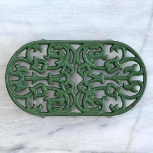 Könnte beinhalten: Ovaler, grüner gusseiserner Untersetzer mit einem kunstvollen, symmetrischen Blumendesign. Der Untersetzer ist in einem matten Grünton lackiert und weist eine Reihe von miteinander verbundenen Blumen- und Scrollmustern auf. Er dient zum Schutz von Oberflächen vor heißem Kochgeschirr.