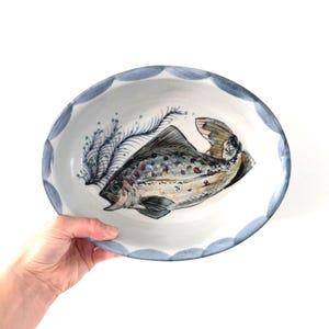 May include: An oval ceramic bowl with a white interior featuring a hand-painted fish illustration with brown, red, and blue details, accompanied by a blue fern-like motif. The rim is decorated with a scalloped pattern in muted blue tones.
