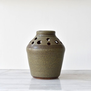 May include: An olive green ceramic vase with a distinctive design. It has circular openings near the top and a darker brown band around the neck. The vase has a rounded body and a slightly flared base, suitable for displaying flowers. The vase is approximately 15 cm tall.